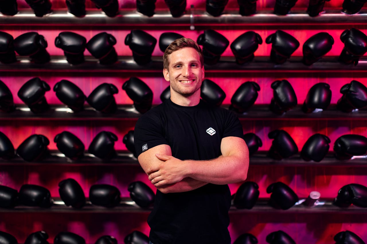 digital A smiling man in a gym stands confidently in front of a wall of boxing gloves.